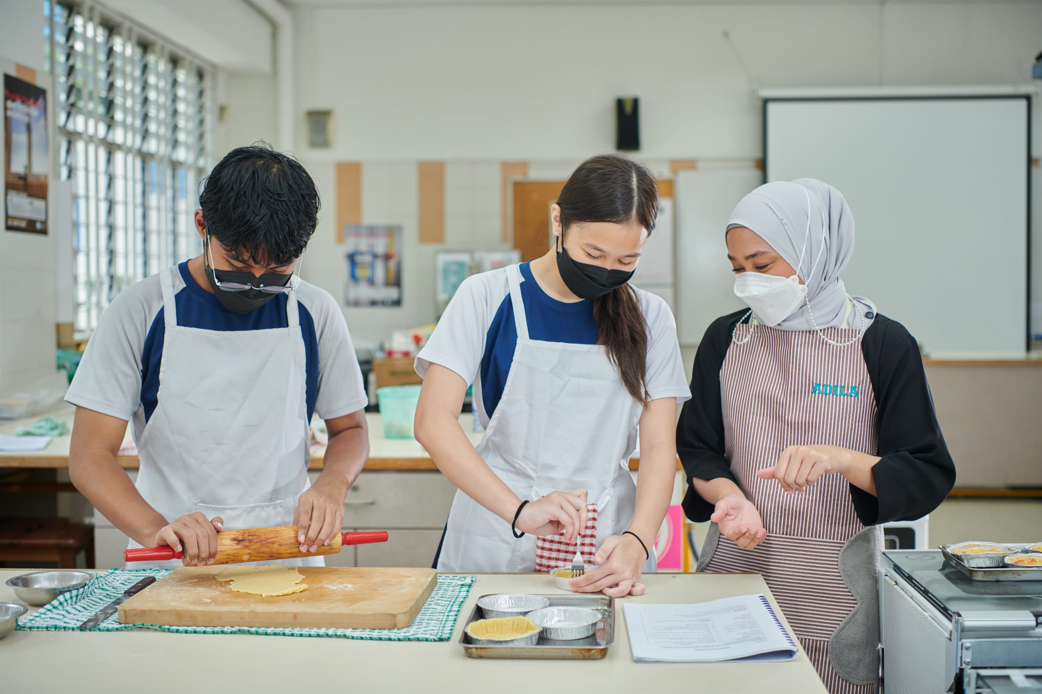 Two masked students and a teacher in aprons work at a kitchen classroom table, rolling dough and filling tart tins.